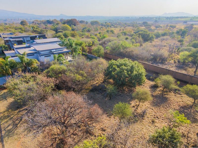 An aerial view of a vacant residential plot featuring natural vegetation, a perimeter wall, and adjacent paved road access in a suburban estate setting.
