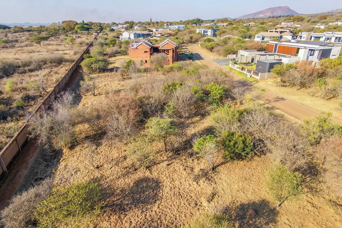 An aerial view of a vacant residential plot featuring natural vegetation, perimeter walling, and established road access within a developed estate.