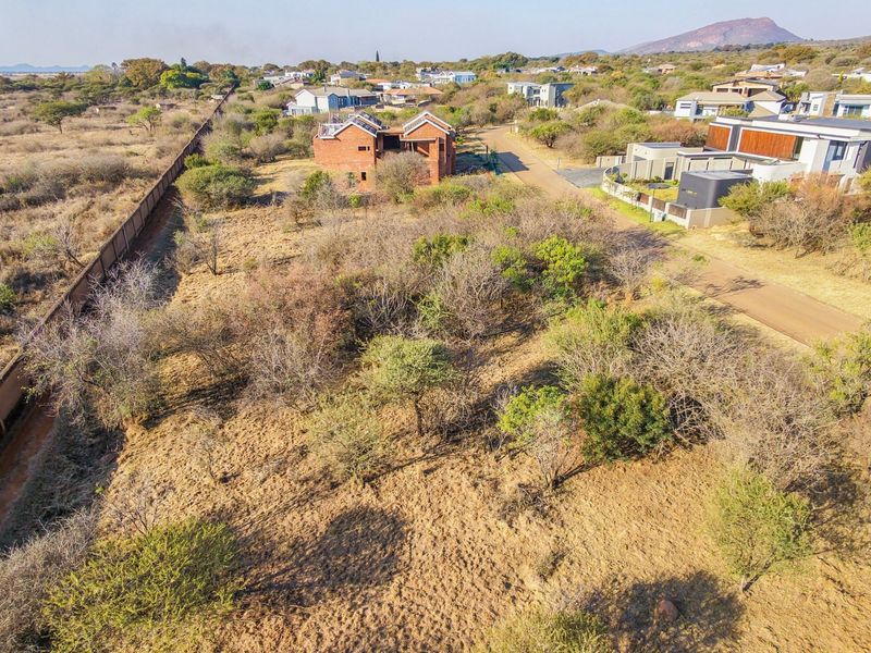 An aerial view of a vacant residential plot featuring natural vegetation, perimeter walling, and established road access within a developed estate.
