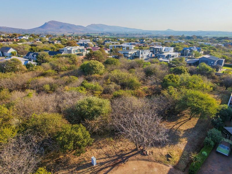 An aerial view of a vacant residential plot featuring natural vegetation, situated within an established estate with surrounding mountain views.