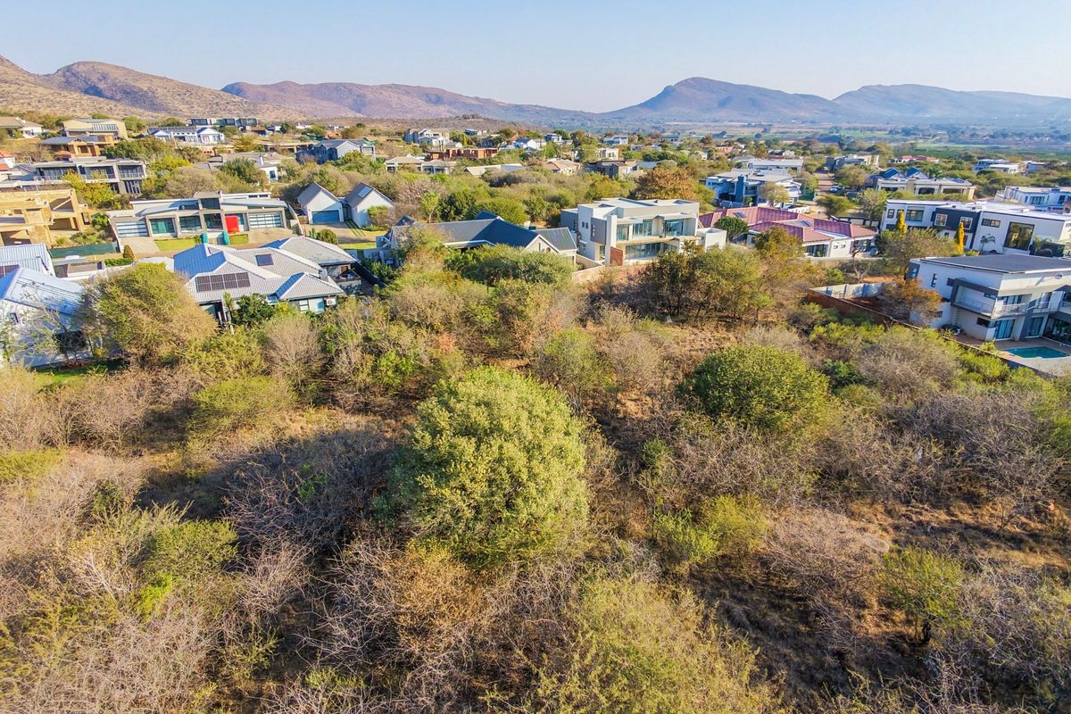 An aerial view of a vacant residential plot surrounded by established modern homes and scenic mountain views in a developed estate.