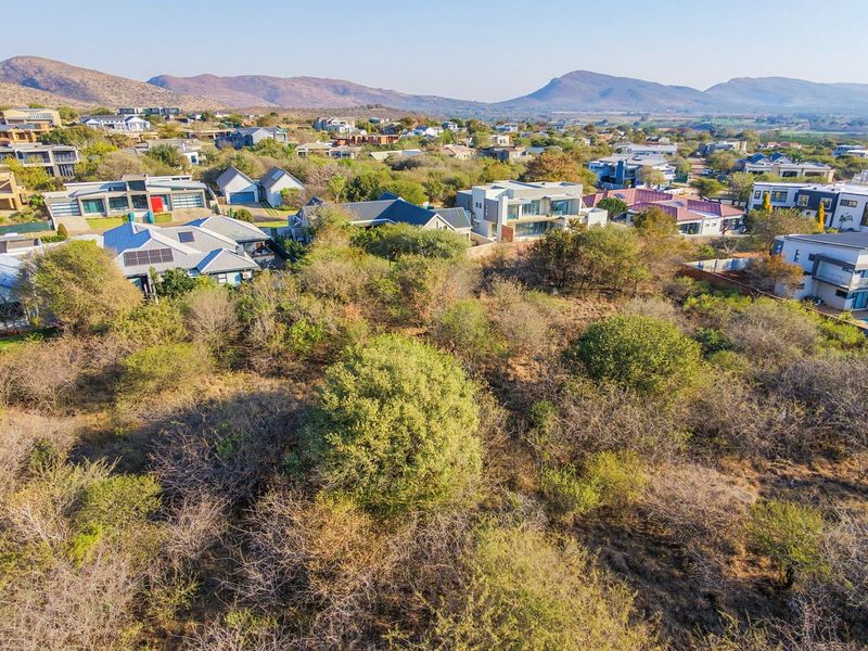 An aerial view of a vacant residential plot surrounded by established modern homes and scenic mountain views in a developed estate.