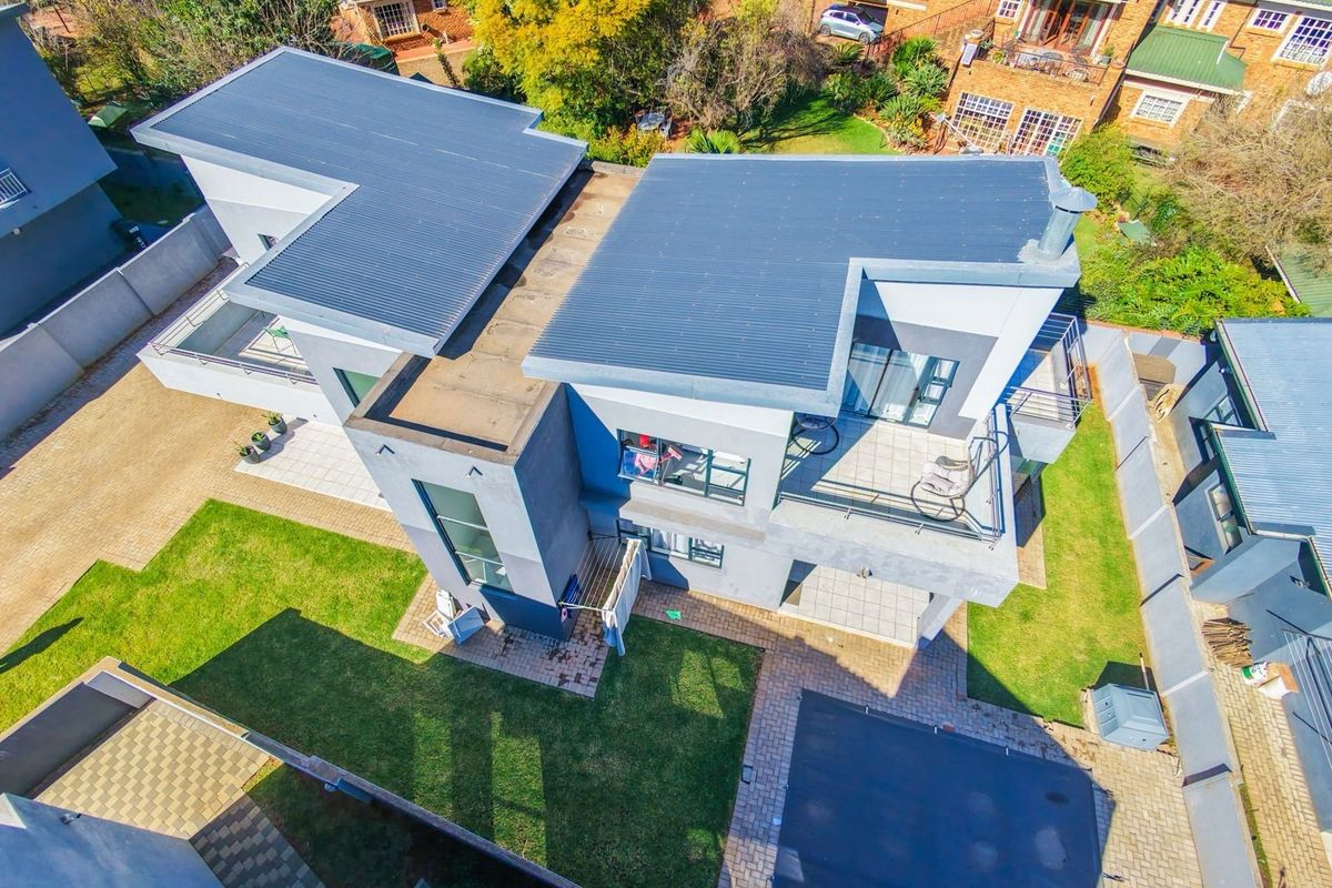 Aerial view of a modern multi-level house featuring a corrugated metal roof, upper-level balcony, paved outdoor areas, and a well-maintained garden.
