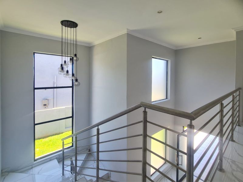 A bright and modern staircase landing featuring polished tile flooring, a sleek stainless steel balustrade, and contemporary pendant lighting with large windows.
