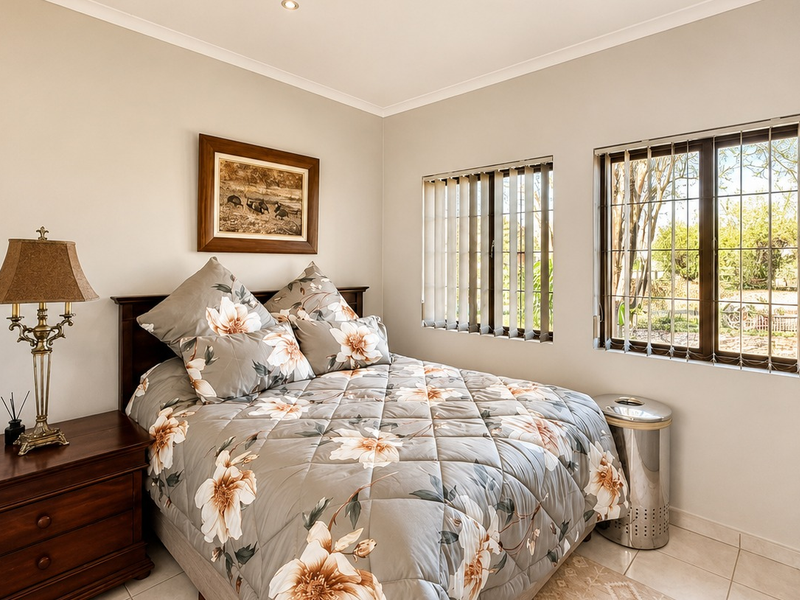 A well-maintained bedroom featuring built-in cupboards, tiled flooring, and large windows with security bars and vertical blinds.