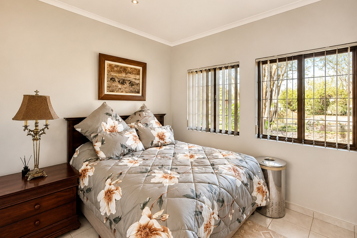 A well-maintained bedroom featuring built-in cupboards, tiled flooring, and large windows with security bars and vertical blinds.