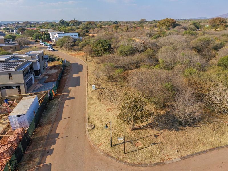 Aerial view of a vacant residential plot situated in a developed estate, featuring established road infrastructure and surrounding natural vegetation.
