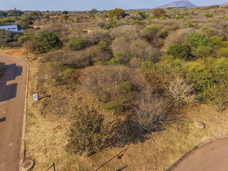 An aerial view of a vacant residential plot featuring natural vegetation, adjacent paved roads, and established utility access points in a suburban estate setting.