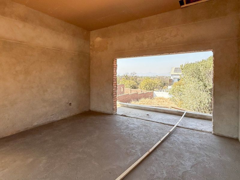 An unfinished bedroom space in shell condition, featuring plastered walls, concrete flooring, and a large open window frame with views of the surrounding area.
