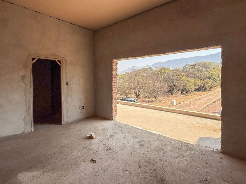 An unfinished bedroom interior in shell condition, featuring plastered walls, concrete flooring, and a large open window frame with views of the surrounding landscape.