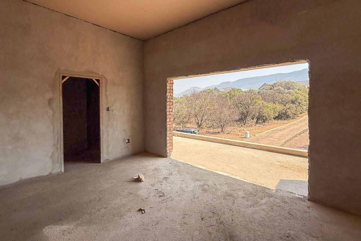 An unfinished bedroom interior in shell condition, featuring plastered walls, concrete flooring, and a large open window frame with views of the surrounding landscape.