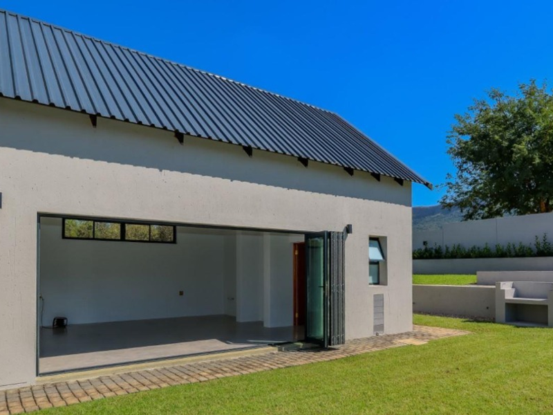 Rear view of a modern structure featuring a metal roof, large bi-fold doors opening to a garden, and a built-in outdoor braai area.