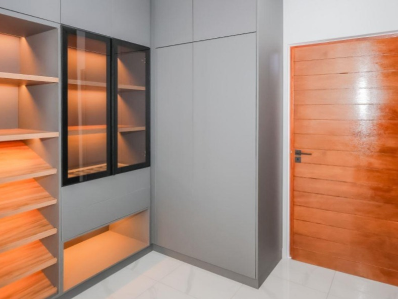 A modern bedroom featuring custom grey built-in cabinetry with integrated shelving, glass display units, warm recessed lighting, and a polished solid wood door.