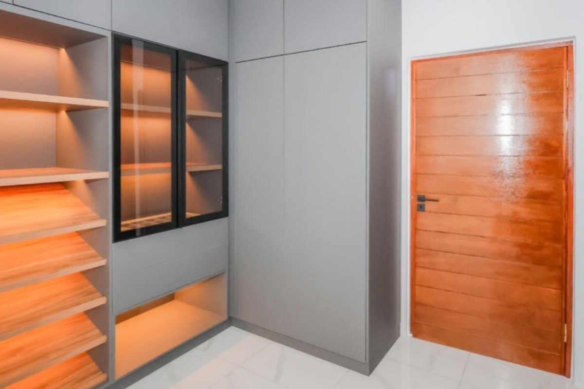 A modern bedroom featuring custom grey built-in cabinetry with integrated shelving, glass display units, warm recessed lighting, and a polished solid wood door.