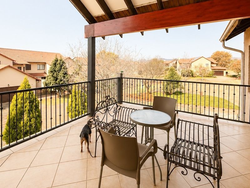 A spacious covered balcony featuring tiled flooring, a decorative wrought iron balustrade, and exposed timber beams, offering pleasant views of the surrounding estate.