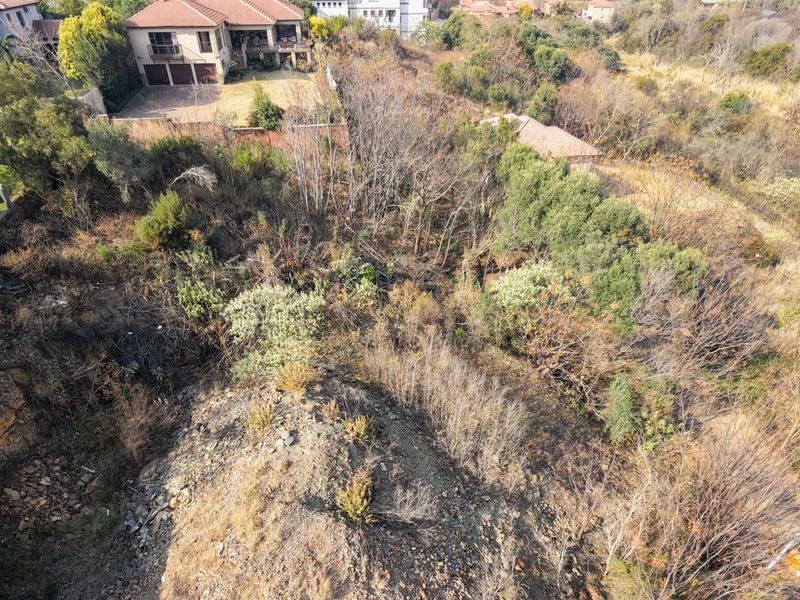 An aerial view of a vacant residential plot featuring natural vegetation, rocky terrain, and surrounding established residential properties.