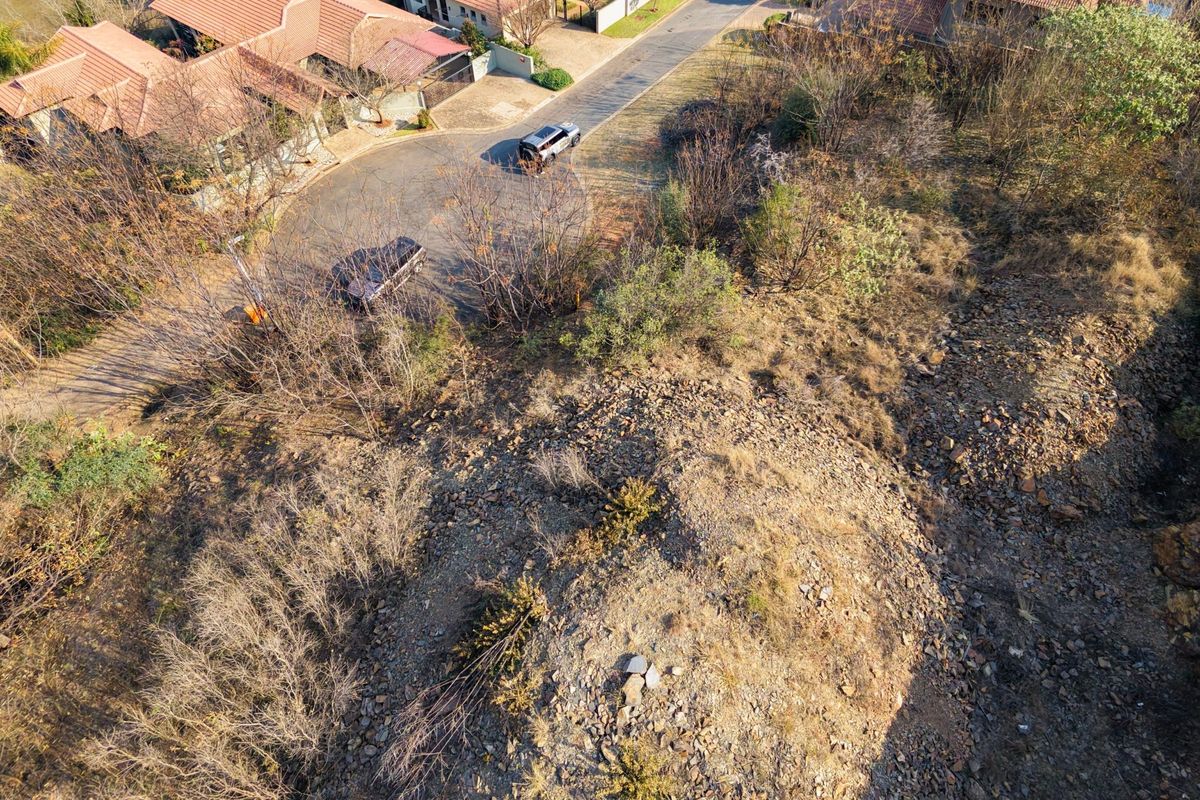 Aerial view of a vacant residential plot featuring natural vegetation and rocky terrain, situated adjacent to an established suburban street.