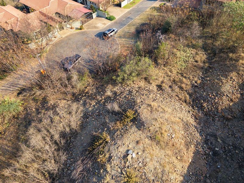 Aerial view of a vacant residential plot featuring natural vegetation and rocky terrain, situated adjacent to an established suburban street.