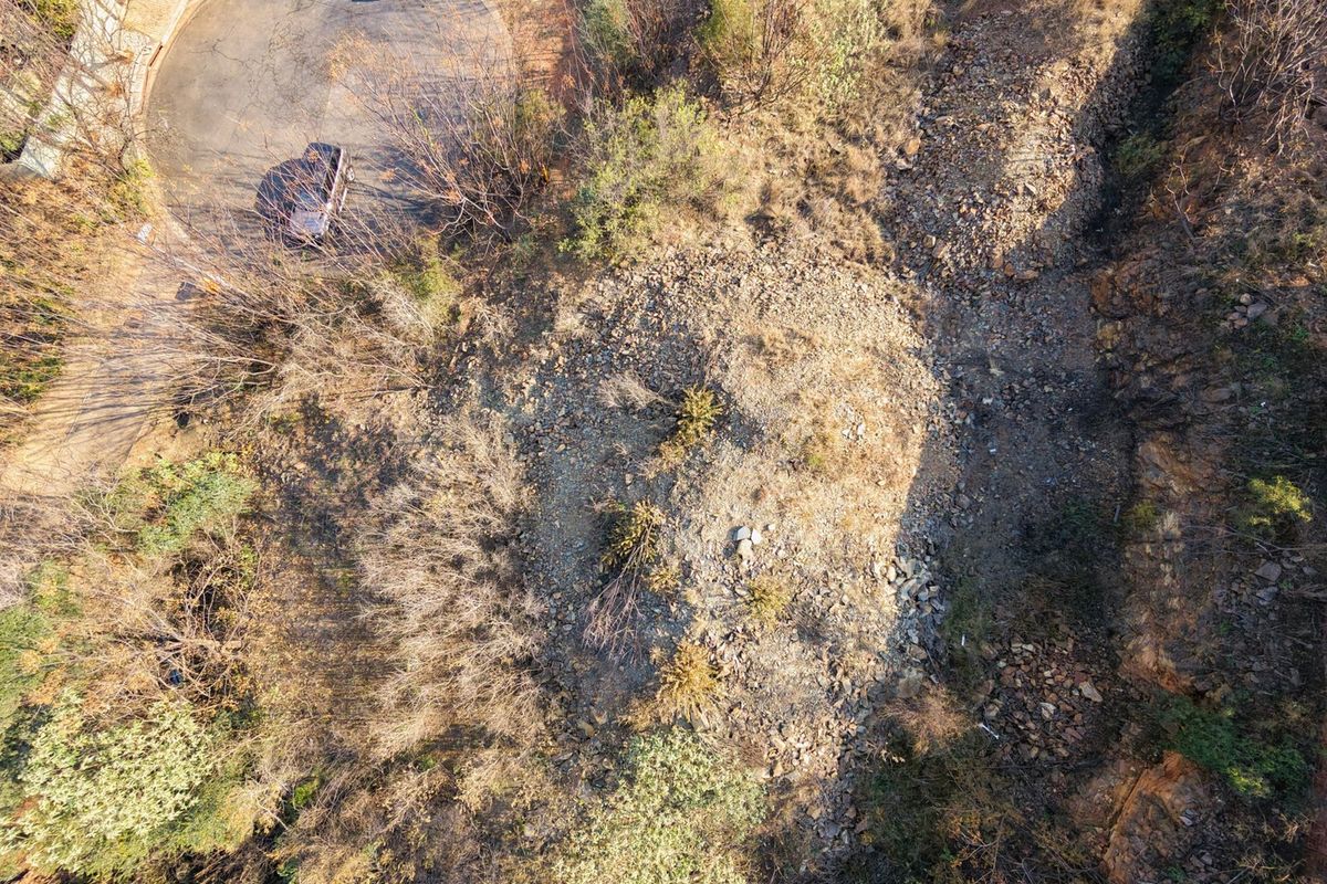 An aerial view of a vacant residential plot featuring natural rocky terrain and established vegetation, adjacent to a paved cul-de-sac.
