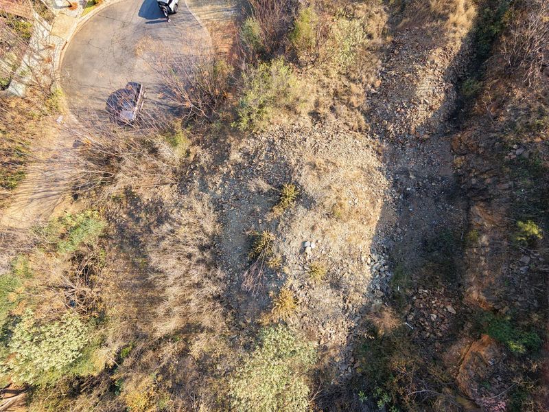 An aerial view of a vacant residential plot featuring natural rocky terrain and established vegetation, adjacent to a paved cul-de-sac.