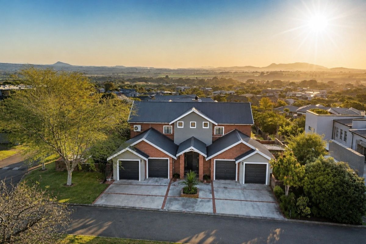 Aerial view of a double-story brick residence featuring a multi-car garage, paved driveway, and a scenic landscape backdrop at sunset.