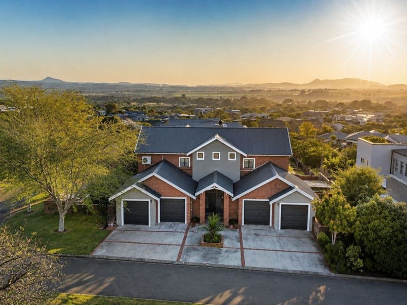 Aerial view of a double-story brick residence featuring a multi-car garage, paved driveway, and a scenic landscape backdrop at sunset.