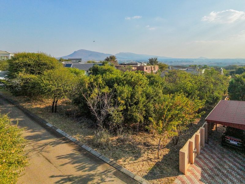Aerial view of a residential vacant plot featuring established trees, a paved driveway, and an adjacent carport structure with mountain views in the background.