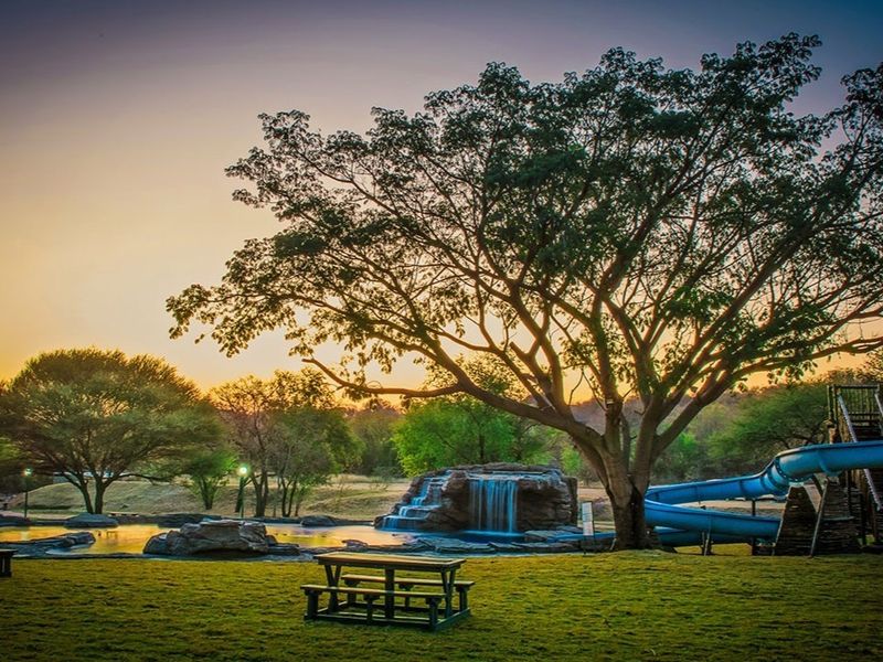 A scenic outdoor pool area featuring a water slide, rock-feature waterfall, and landscaped grounds at sunset.