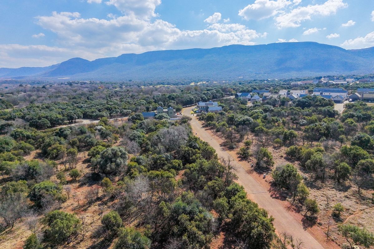 An aerial view of a residential estate featuring unpaved roads, native bushveld vegetation, and scenic mountain views in the background.