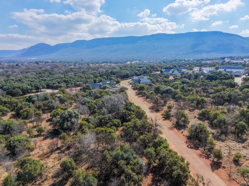 An aerial view of a residential estate featuring unpaved roads, native bushveld vegetation, and scenic mountain views in the background.