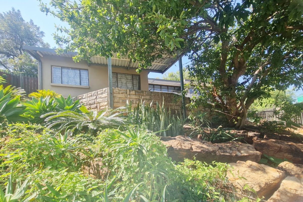 A view of the rear exterior featuring a stone retaining wall, a covered patio area, and a mature landscaped garden.