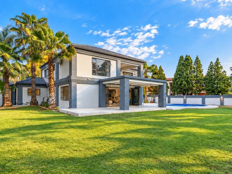 A well-maintained rear exterior view of a two-story house featuring a covered patio, balcony, swimming pool, and expansive landscaped garden.