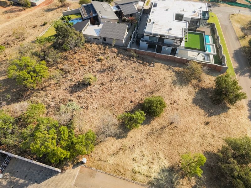 An aerial view of a vacant residential plot featuring native vegetation, situated in a developed estate with neighboring modern homes.
