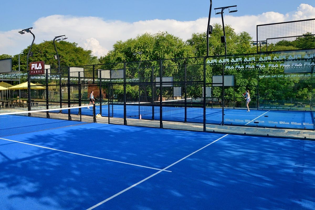 A well-maintained outdoor padel court featuring blue synthetic turf, professional glass paneling, perimeter fencing, and integrated floodlighting for evening play.