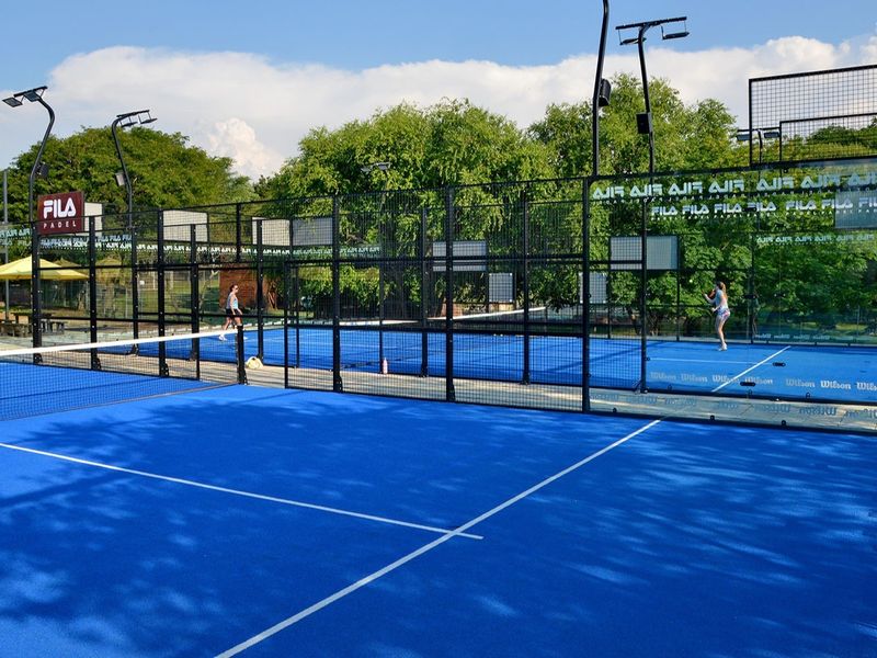 A well-maintained outdoor padel court featuring blue synthetic turf, professional glass paneling, perimeter fencing, and integrated floodlighting for evening play.