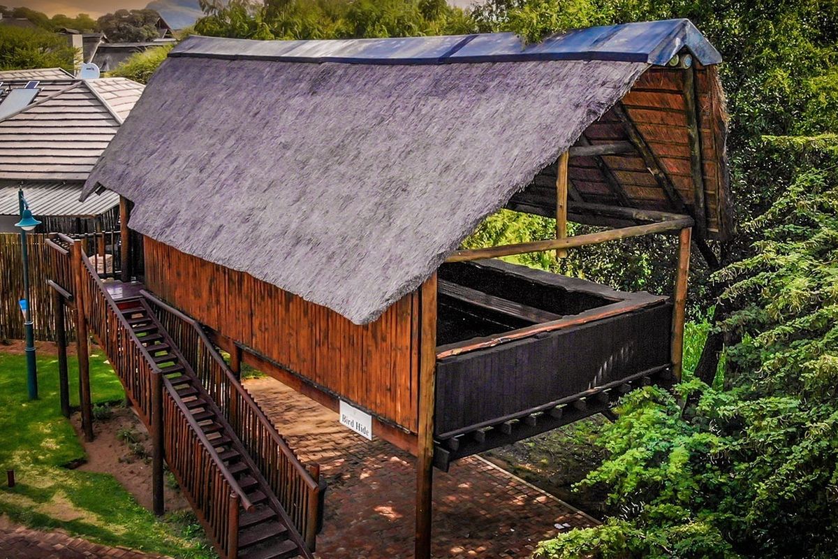 An elevated timber bird hide structure featuring a traditional thatched roof, accessible via a wooden staircase, situated within a landscaped residential garden setting.