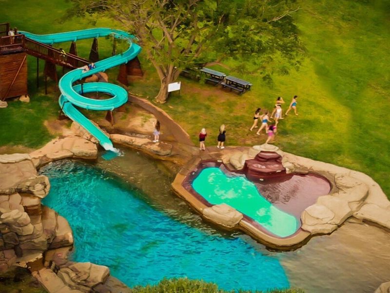 Aerial view of a recreational pool area featuring a water slide, rock-scaped swimming pools, and surrounding grass lawn.