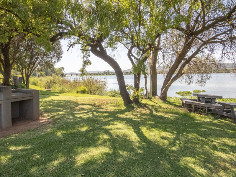 A scenic waterfront garden area featuring a built-in braai, mature trees, and expansive lawn space overlooking the lake.