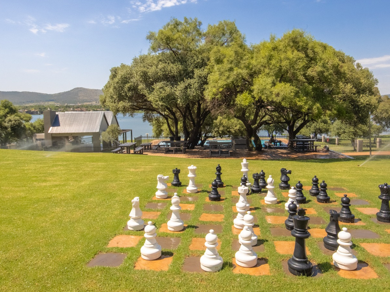 A well-maintained garden area featuring a large outdoor chess set, mature trees, and a scenic view of the nearby lake and clubhouse facilities.