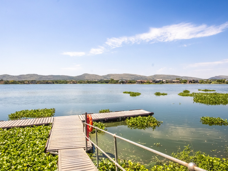 A scenic view of a calm lake featuring a floating jetty with a metal handrail and lifebuoy, surrounded by natural aquatic vegetation.