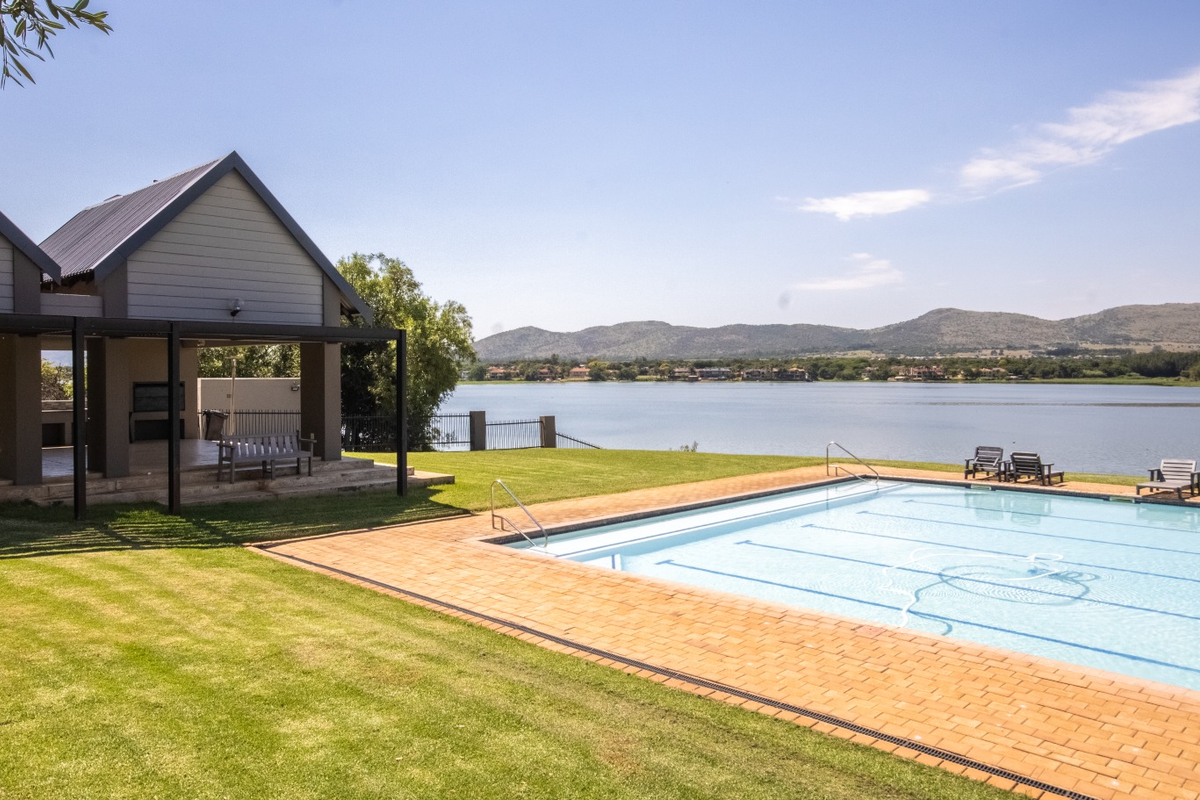 A well-maintained swimming pool area featuring brick coping, a spacious lawn, and a covered patio structure overlooking a scenic lake and mountain view.