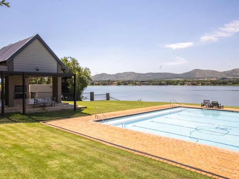 A well-maintained swimming pool area featuring brick coping, a spacious lawn, and a covered patio structure overlooking a scenic lake and mountain view.