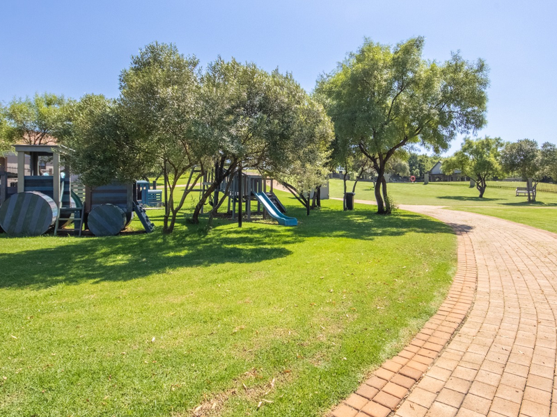 A well-maintained communal garden area featuring a paved walkway, expansive green lawns, mature trees, and a children's playground structure.