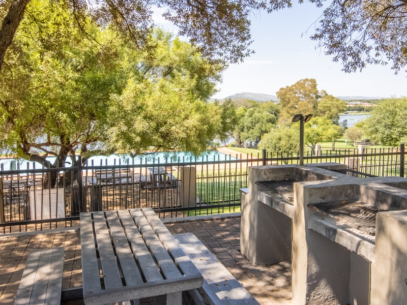 Outdoor entertainment area featuring a built-in braai, paved flooring, and metal fencing, overlooking a scenic landscape with mature trees.