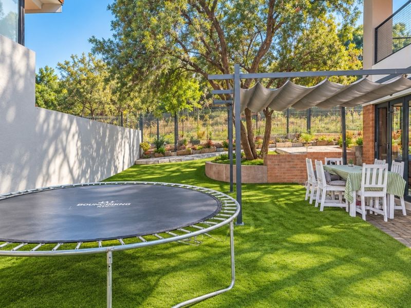 A well-maintained rear garden featuring a paved patio area with a retractable pergola, artificial lawn, and tiered landscaping with a retaining wall.