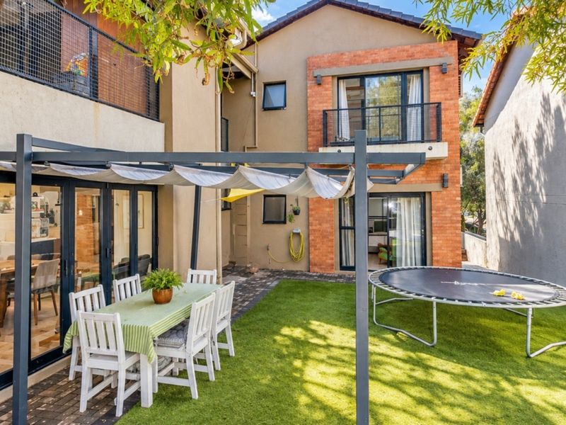Rear exterior view featuring a paved patio with a retractable pergola, artificial lawn, and a multi-story brick and plaster facade with balcony access.