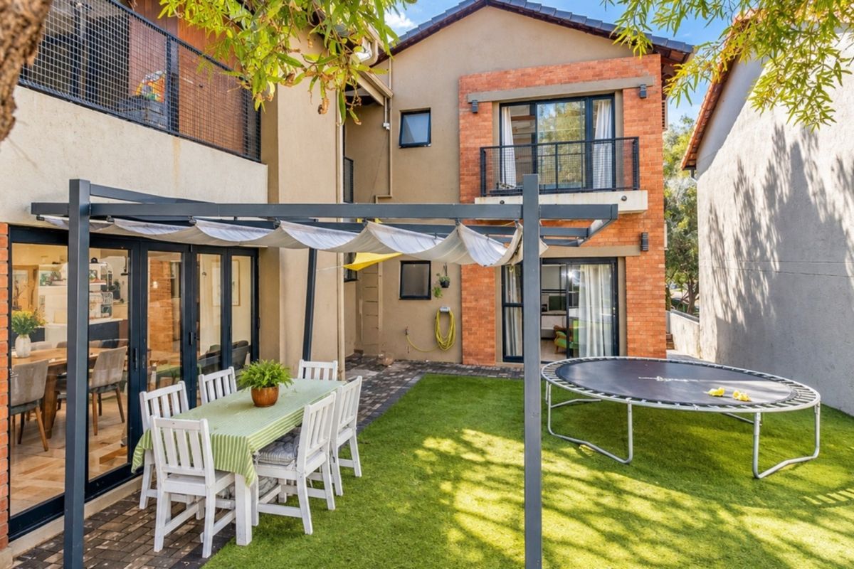 Rear exterior view featuring a paved patio with a retractable pergola, artificial lawn, and a multi-story brick and plaster facade with balcony access.