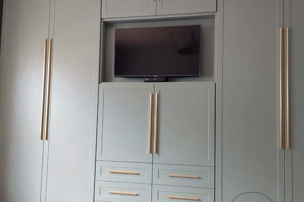 A modern bedroom featuring custom floor-to-ceiling built-in cabinetry in a soft sage green finish with elegant brass hardware and integrated media storage.