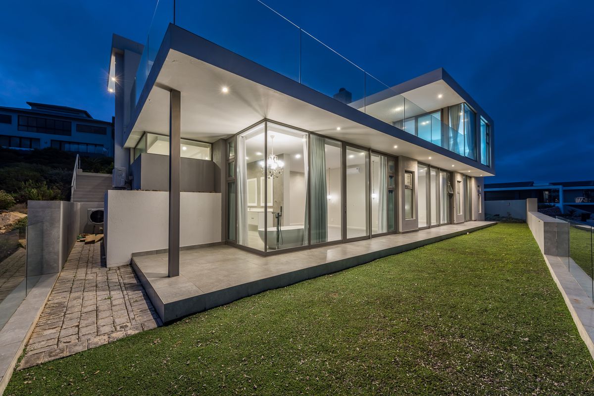 Modern architectural rear elevation featuring expansive glass sliding doors, a covered tiled patio, and a glass-railed balcony overlooking the landscaped garden at twilight.