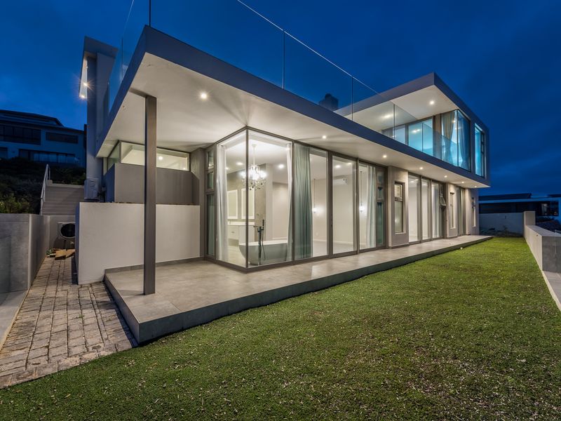 Modern architectural rear elevation featuring expansive glass sliding doors, a covered tiled patio, and a glass-railed balcony overlooking the landscaped garden at twilight.